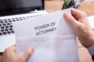 A Person's Hand Holding Power Of Attorney Document In White Envelope In Front Of Laptop