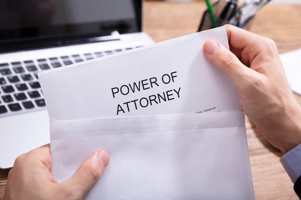 A Person's Hand Holding Power Of Attorney Document In White Envelope In Front Of Laptop