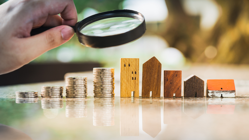 Hand holding magnifying glass and looking at house model with row of coin money, house selection, real estate concept.